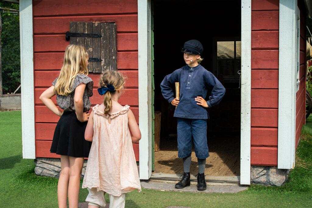 A boy dressed as Emil i Lönneberga in front of a red cabin. Two girls are looking at him.