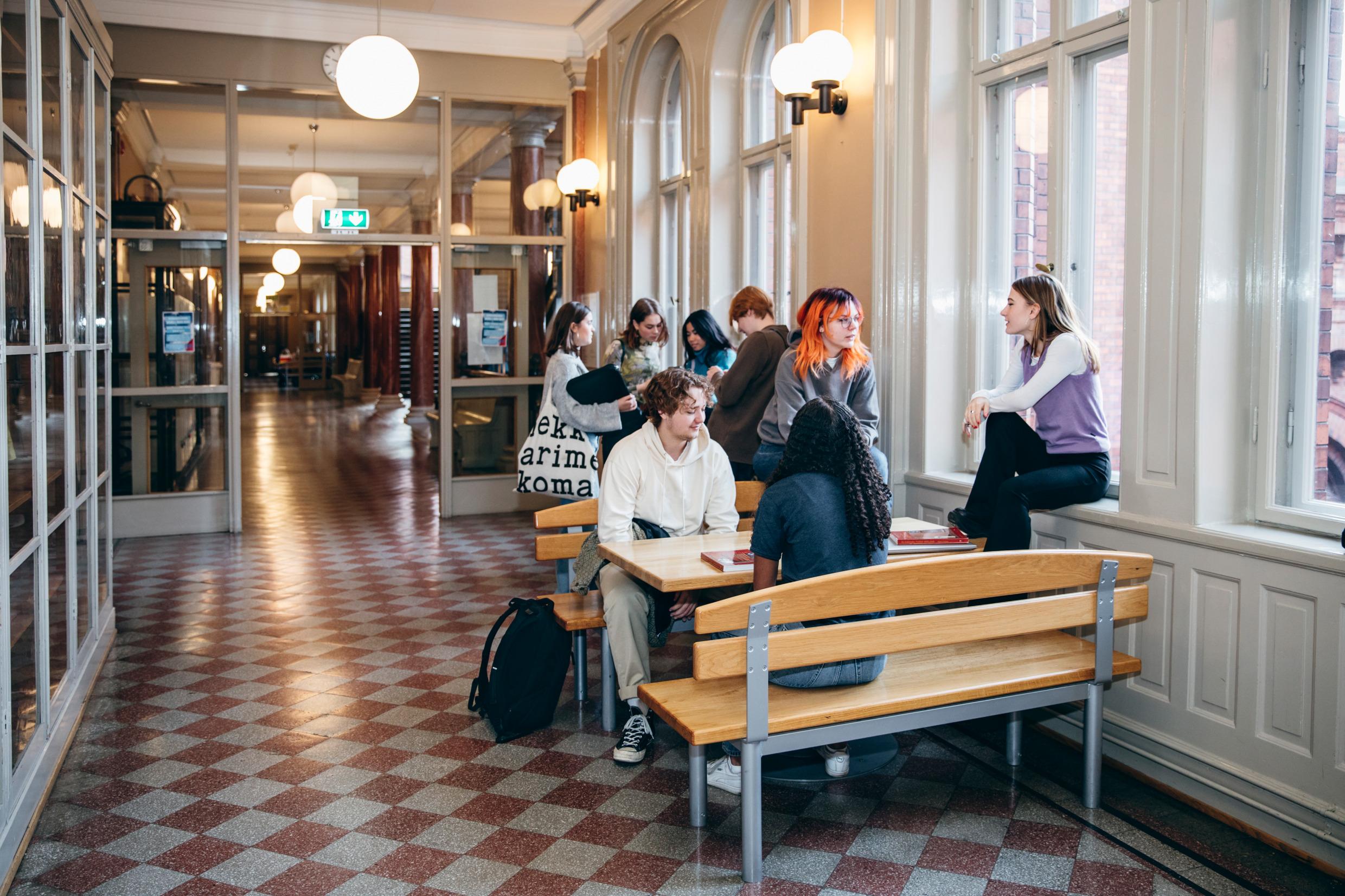 Teenagers sit together by tables in a long hallway.