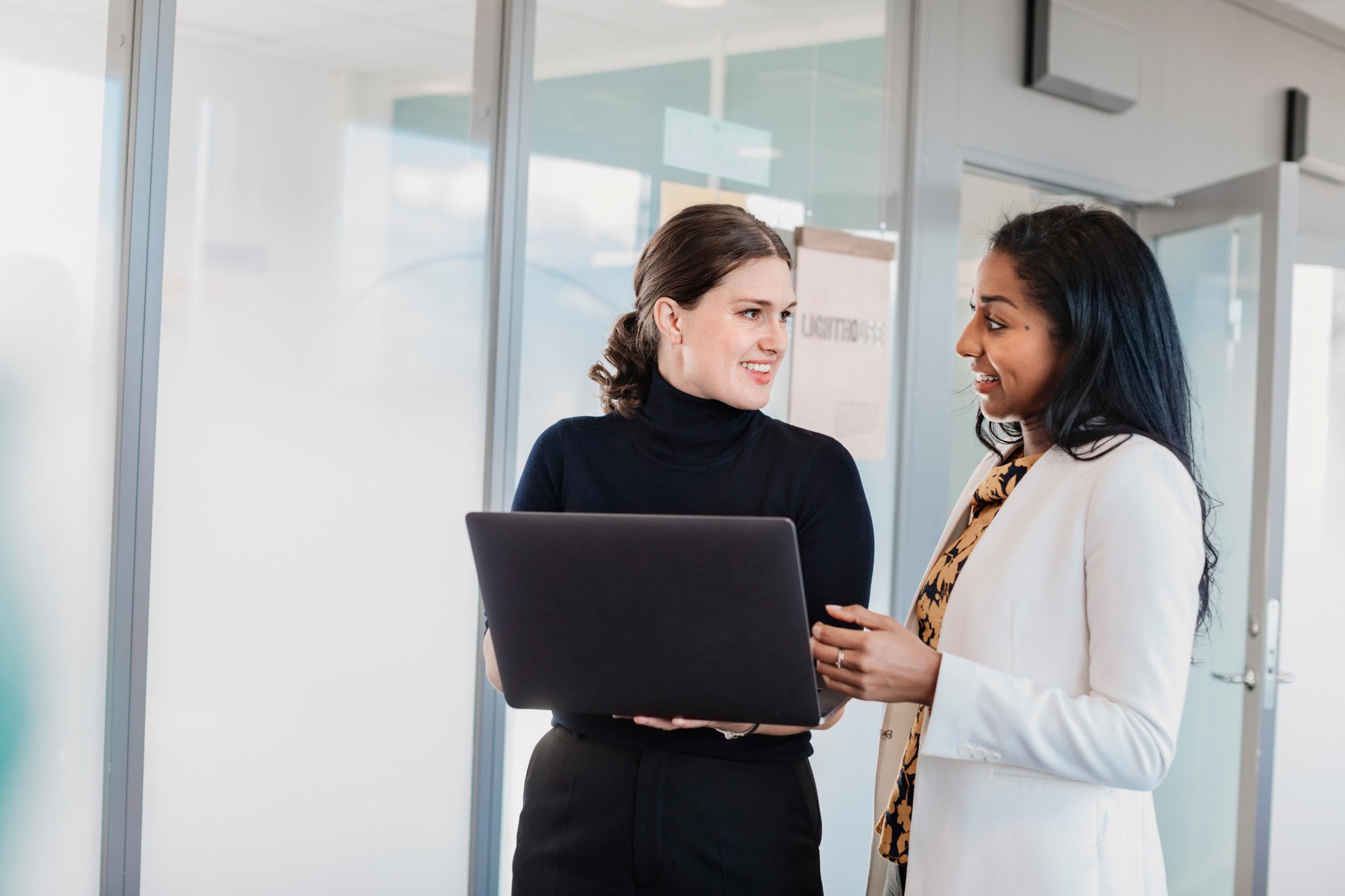 Two women stand smiling, looking at each other. One of them holds a laptop.