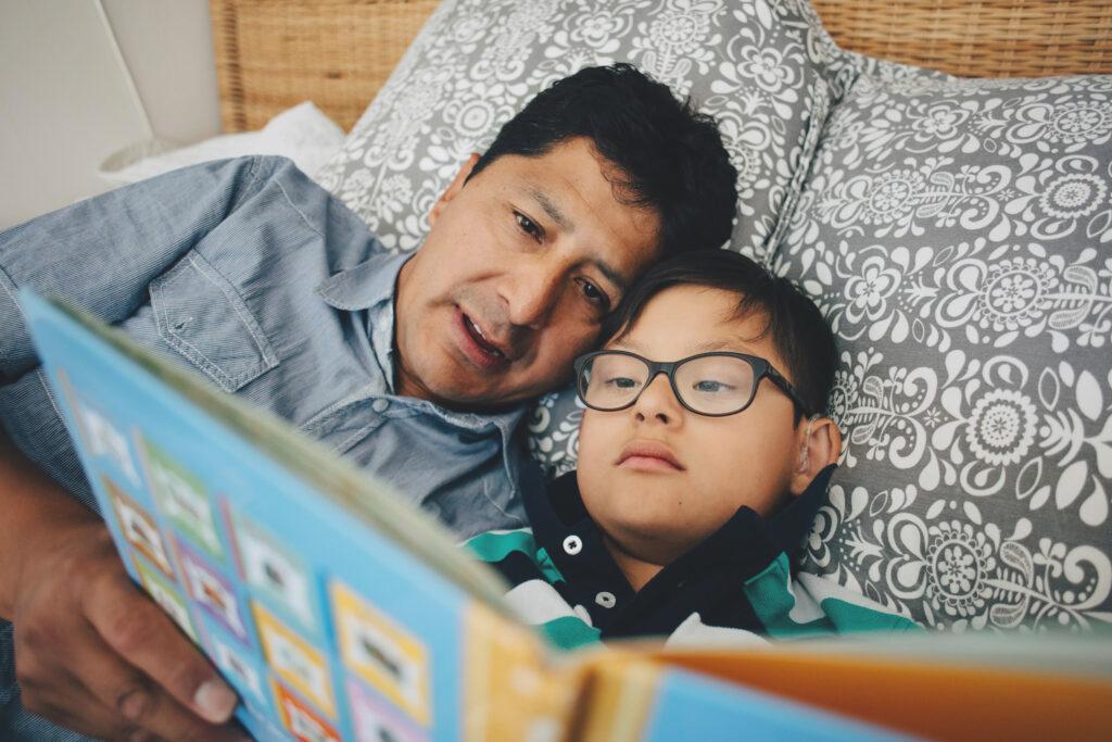 A man and a boy is lying in bed, reading from a book.