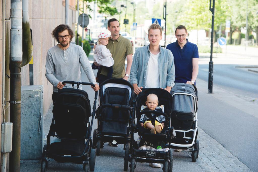 Four men with baby strollers walk down the street. Each man has a baby stroller.