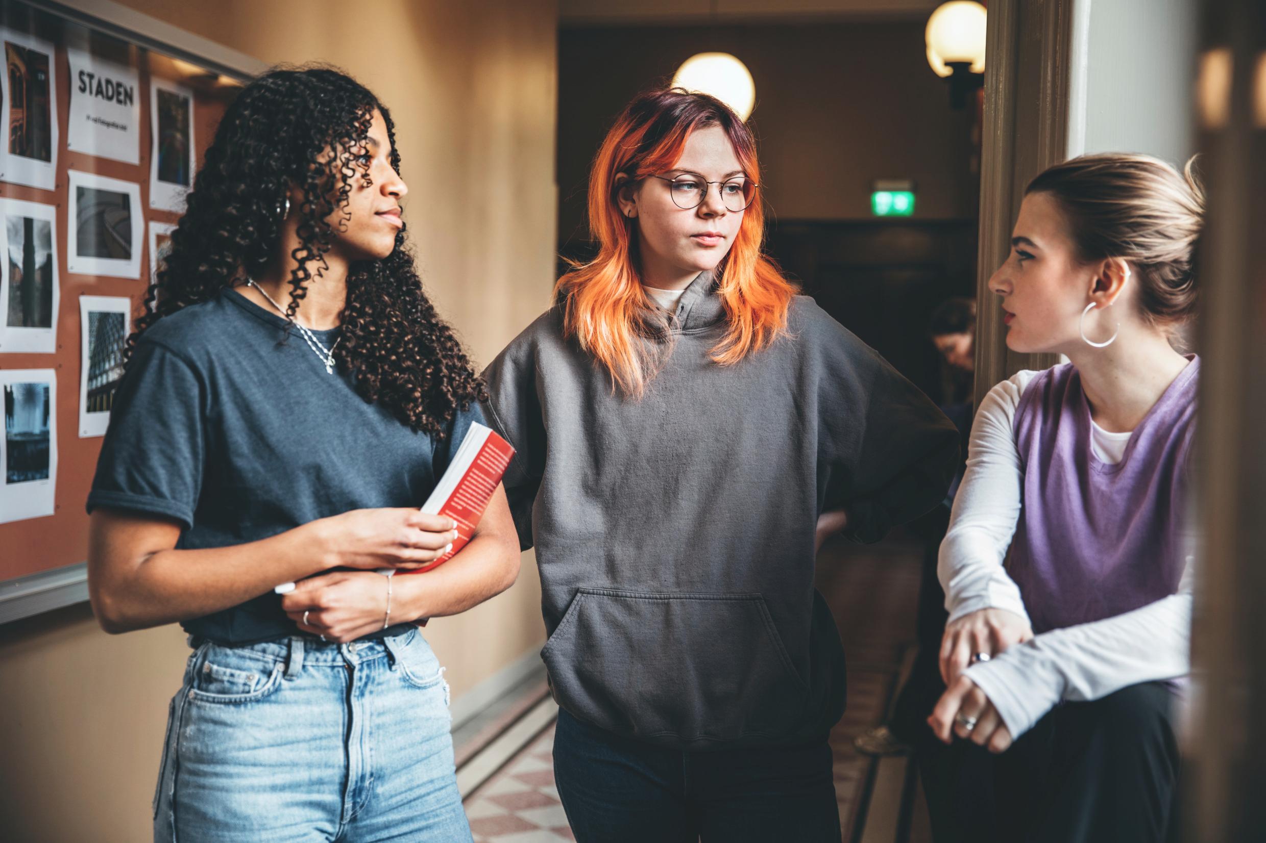 Three teenage girls stand in a hallway, talking.