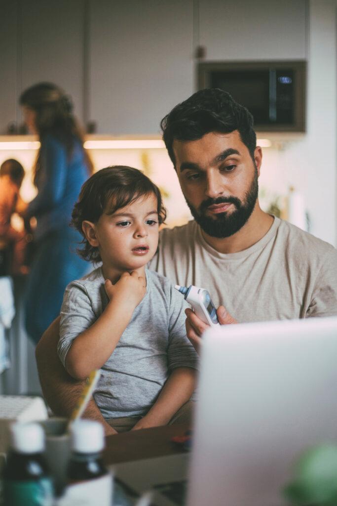 A father sits with a sick child in his lap in front of the computer and holds a thermometer.