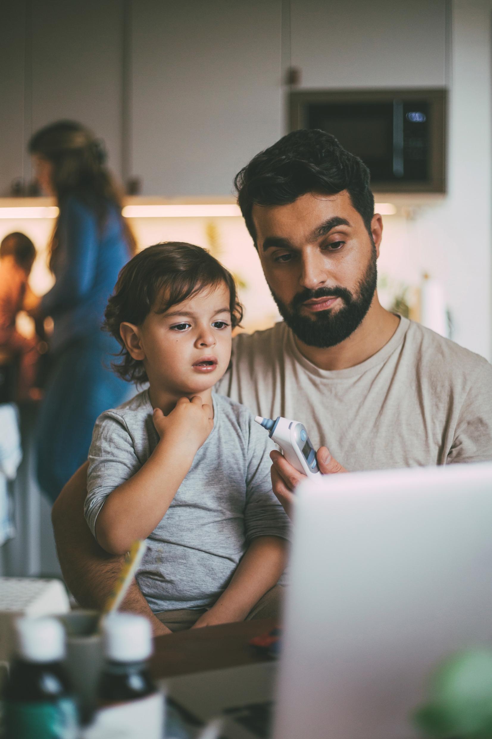 A father sits with a sick child in his lap in front of the computer and holds a thermometer.