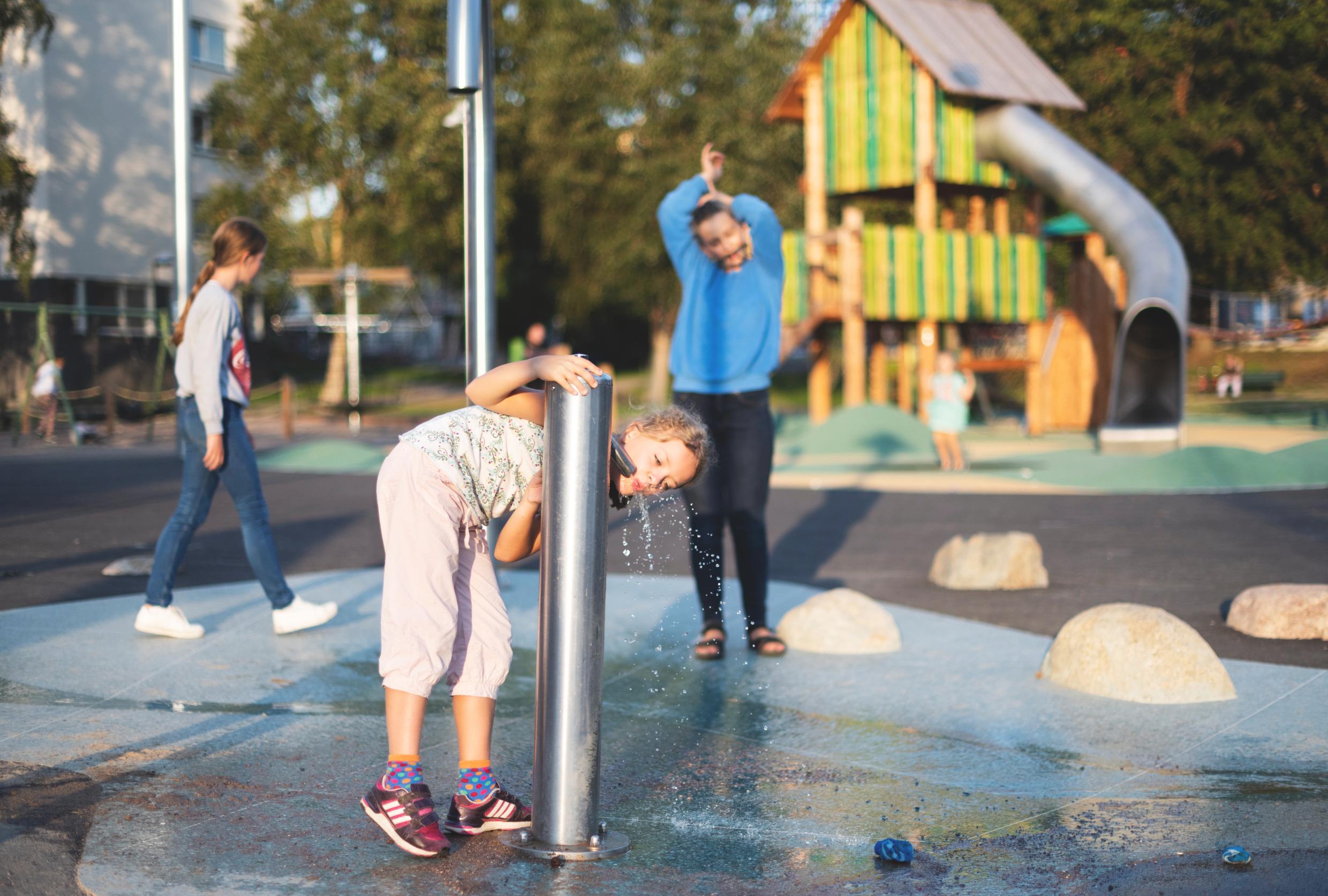 A girl is drinking from a water fountain at a playground.