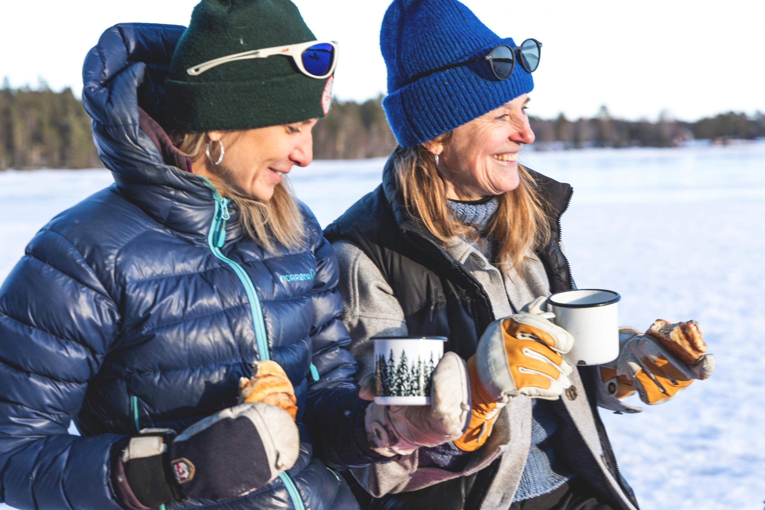 Two women are each holding a coffee cup and a sandwich. They are smiling.