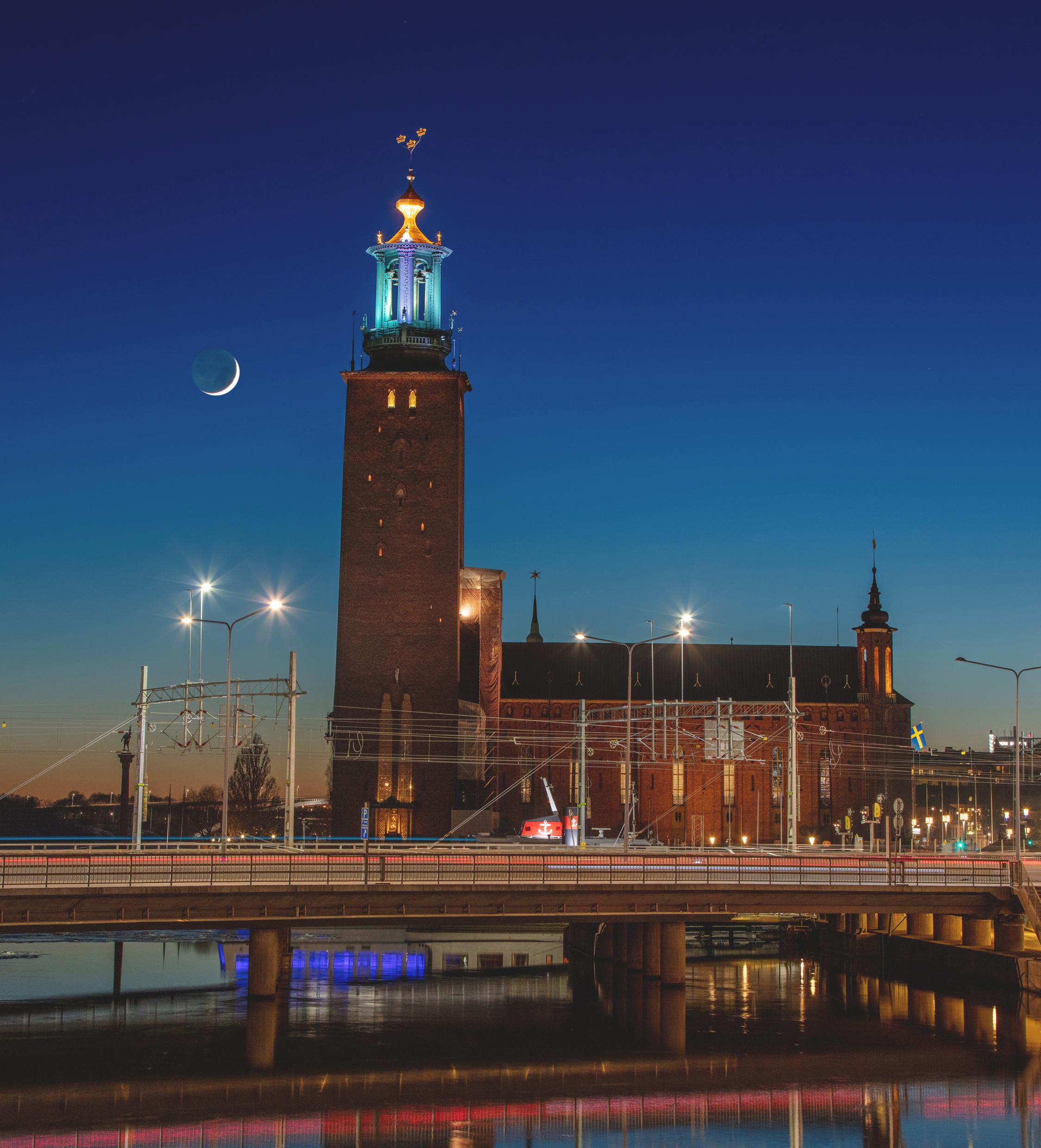 The new moon shines behind a large brick building with a huge clock tower.