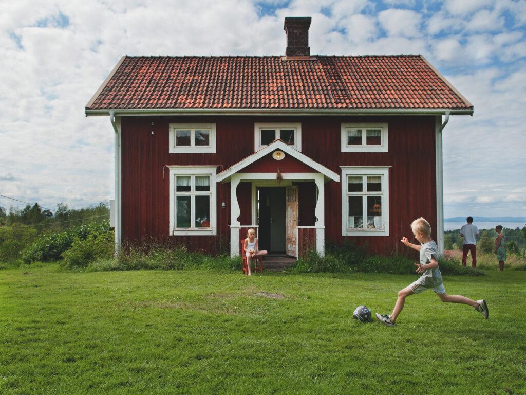 A family vacation setting outside a red house with white borders. A boy is kicking a ball; a girl sits on a chair, and the parents stand next to the house.
