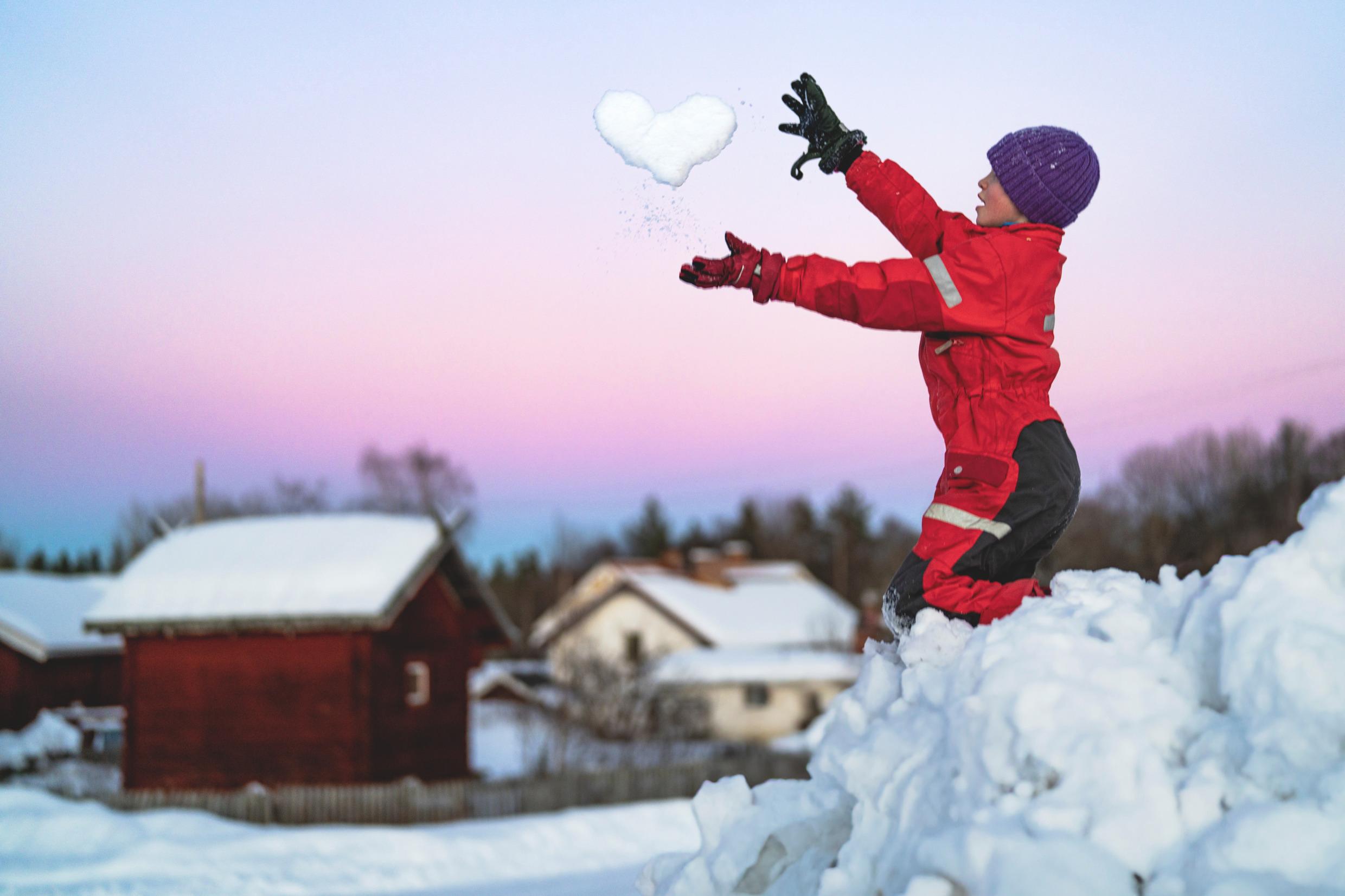 A child is playing in the snow and is throwing snow shaped like a heart in the air.