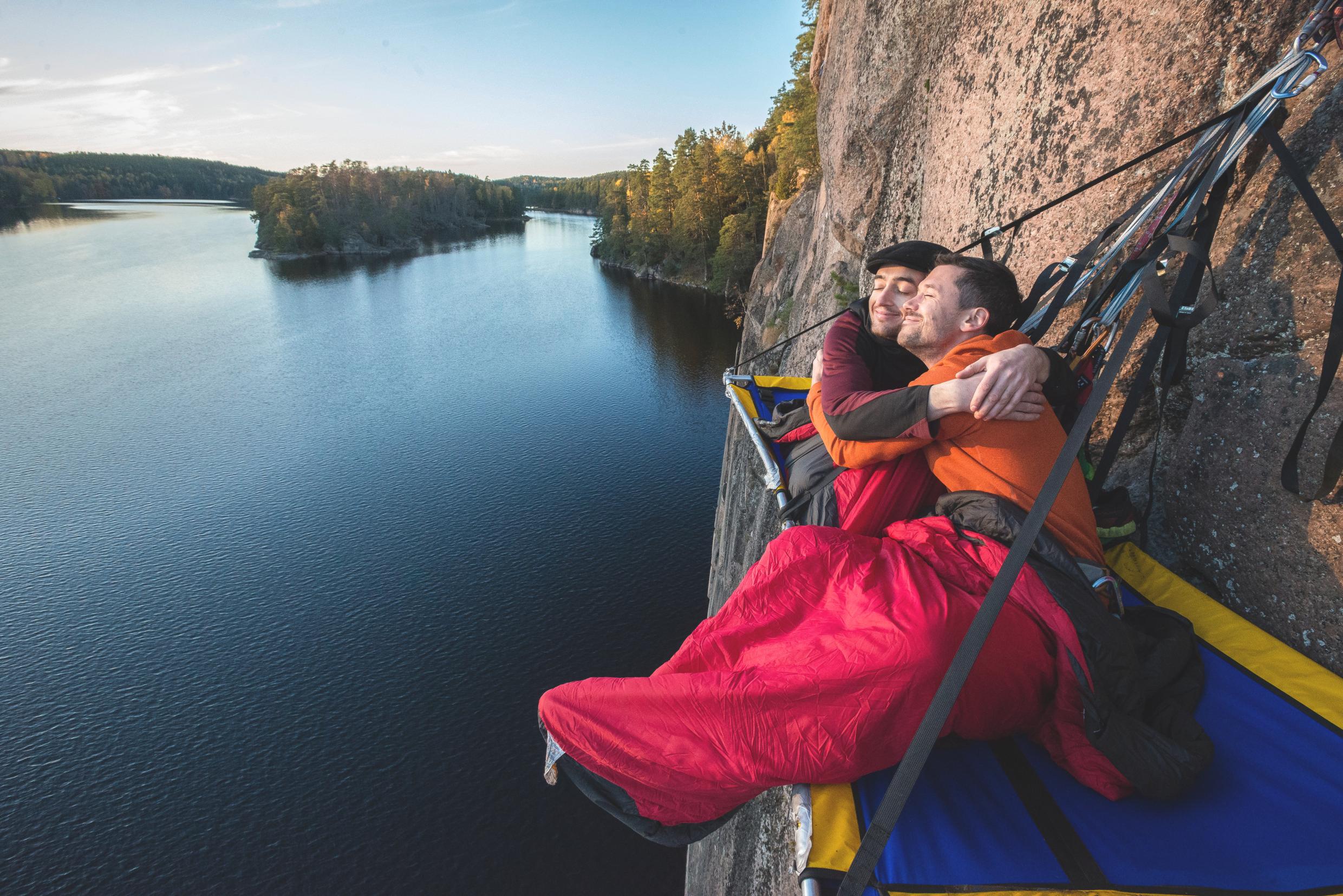 Two men getting ready for sleep while suspended on a cliffside over the water.