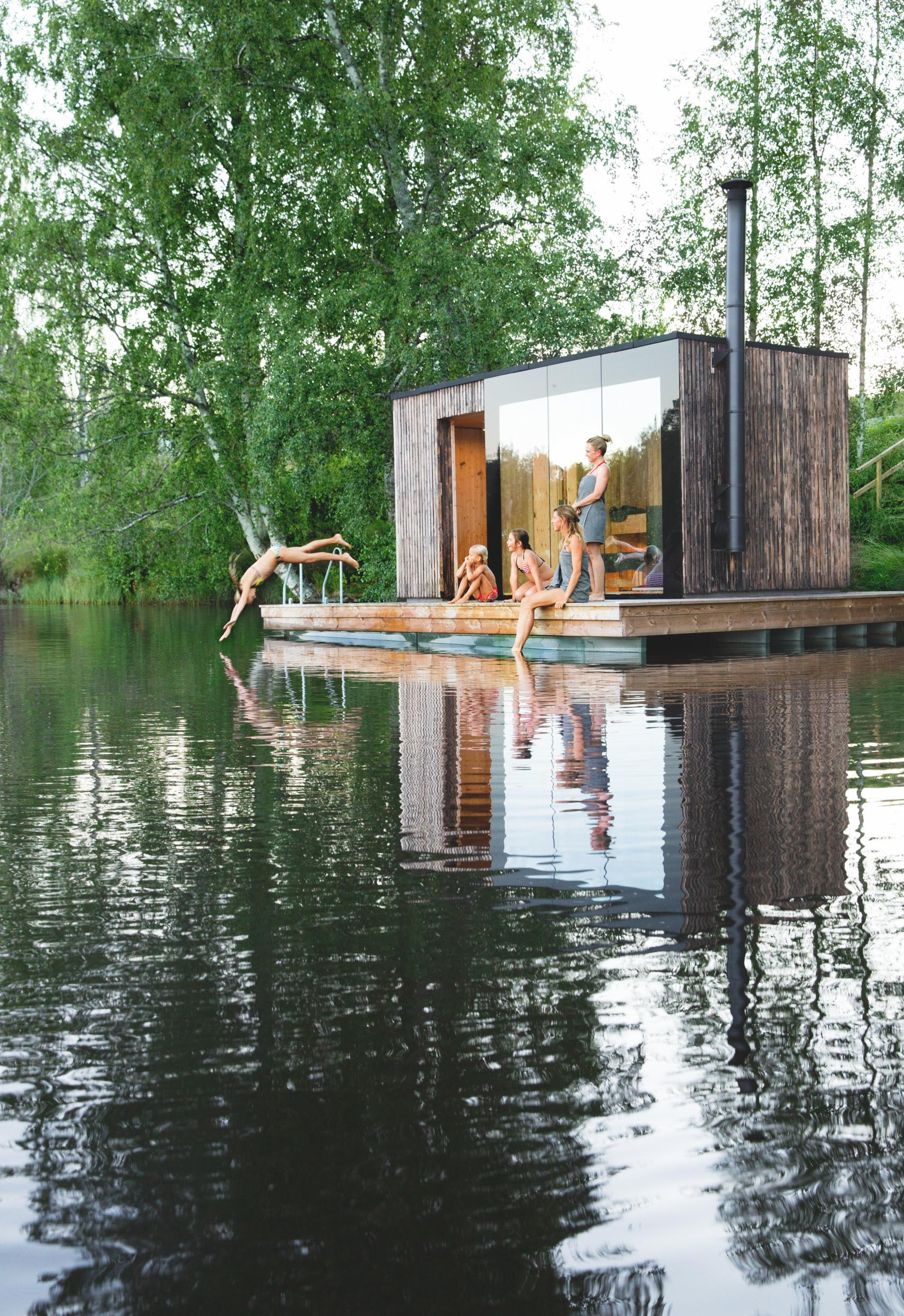 Agroup of women and girls in and around a sauna built on a small wooden pier by a lake.