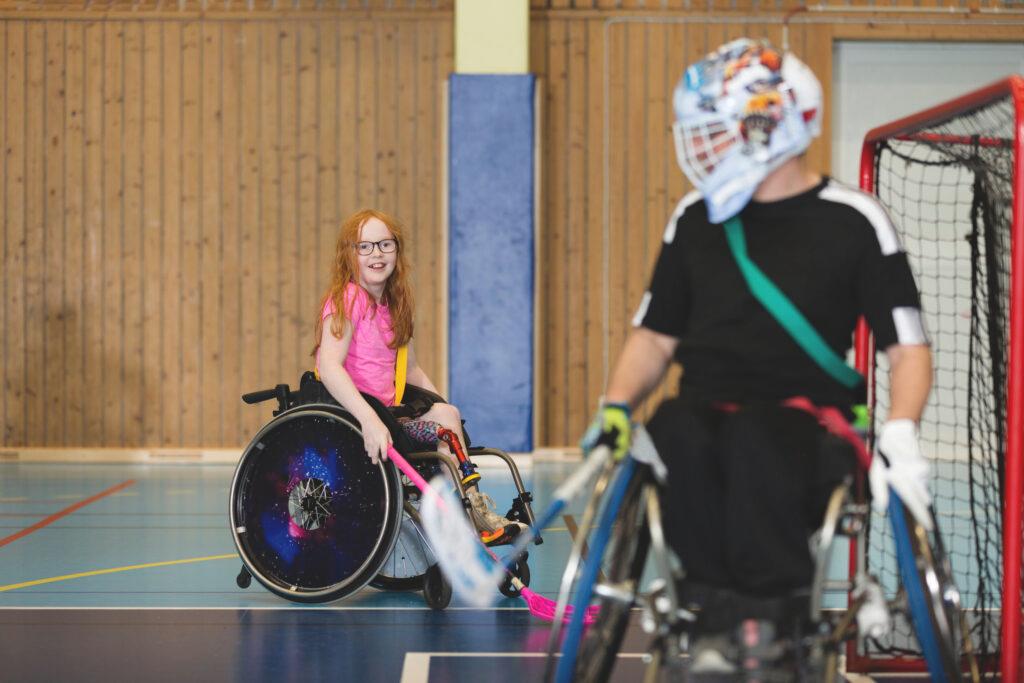 A girl plays wheelchair floorball, in the foreground a male goalkeeper in a wheelchair.