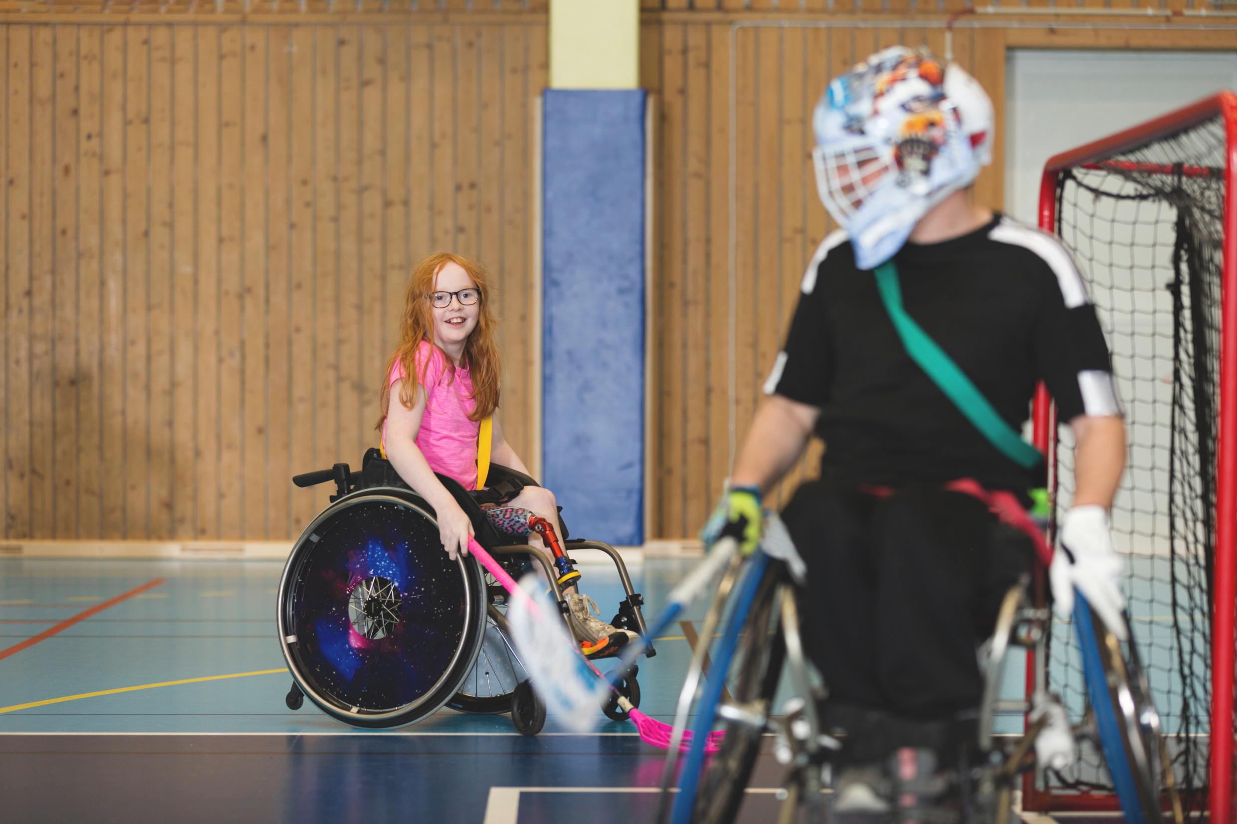 A girl plays wheelchair floorball, in the foreground a male goalkeeper in a wheelchair.
