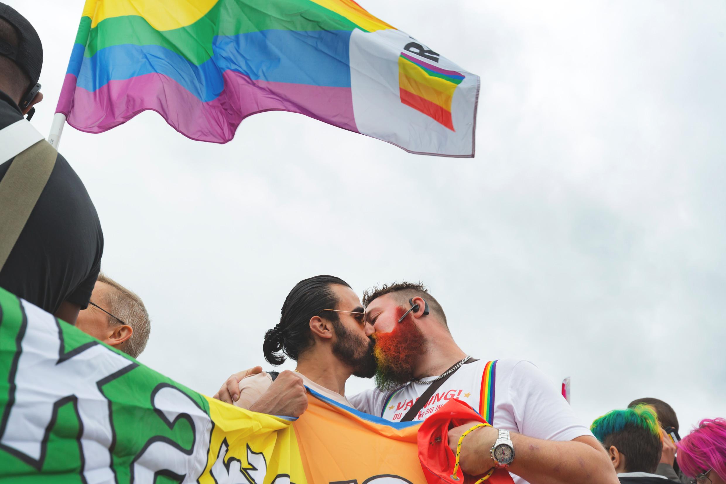 Two men kiss under a rainbow flag.