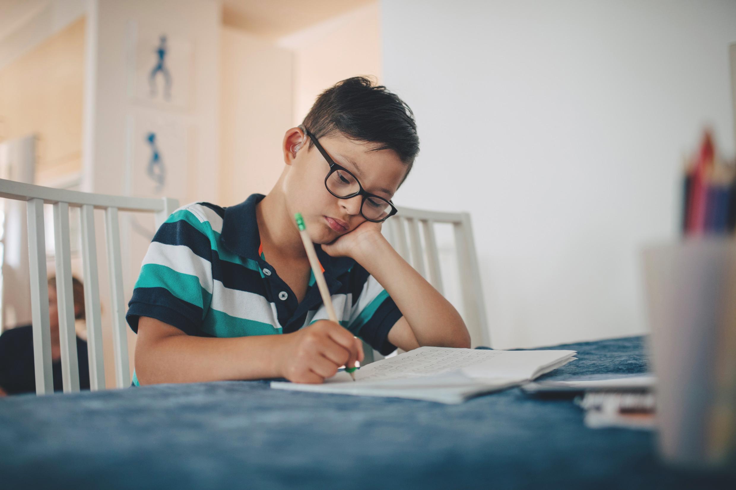 A boy sits by a table, writing in a notebook.