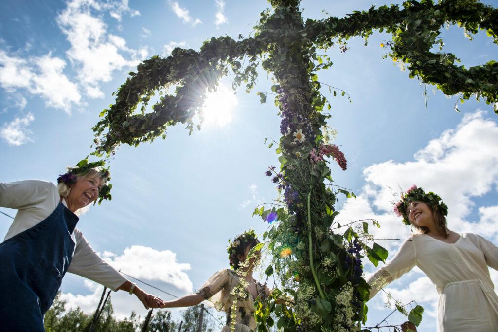 Three women dance around a maypole. The sky is blue and the sun is shining.
