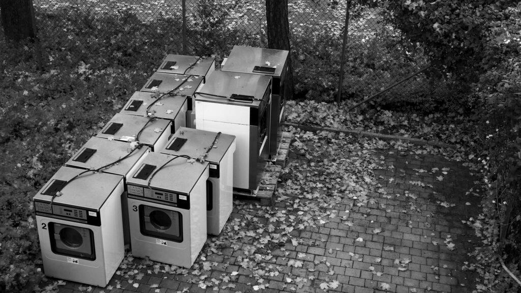 Black and white photo of old washing machines in a backyard filled with autumn leaves.