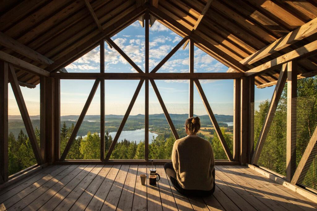 A woman sits on the floor of an open loft in the morning light, with freshly brewed coffee at her side, overlooking the mountains and the forest in Hälsingland.