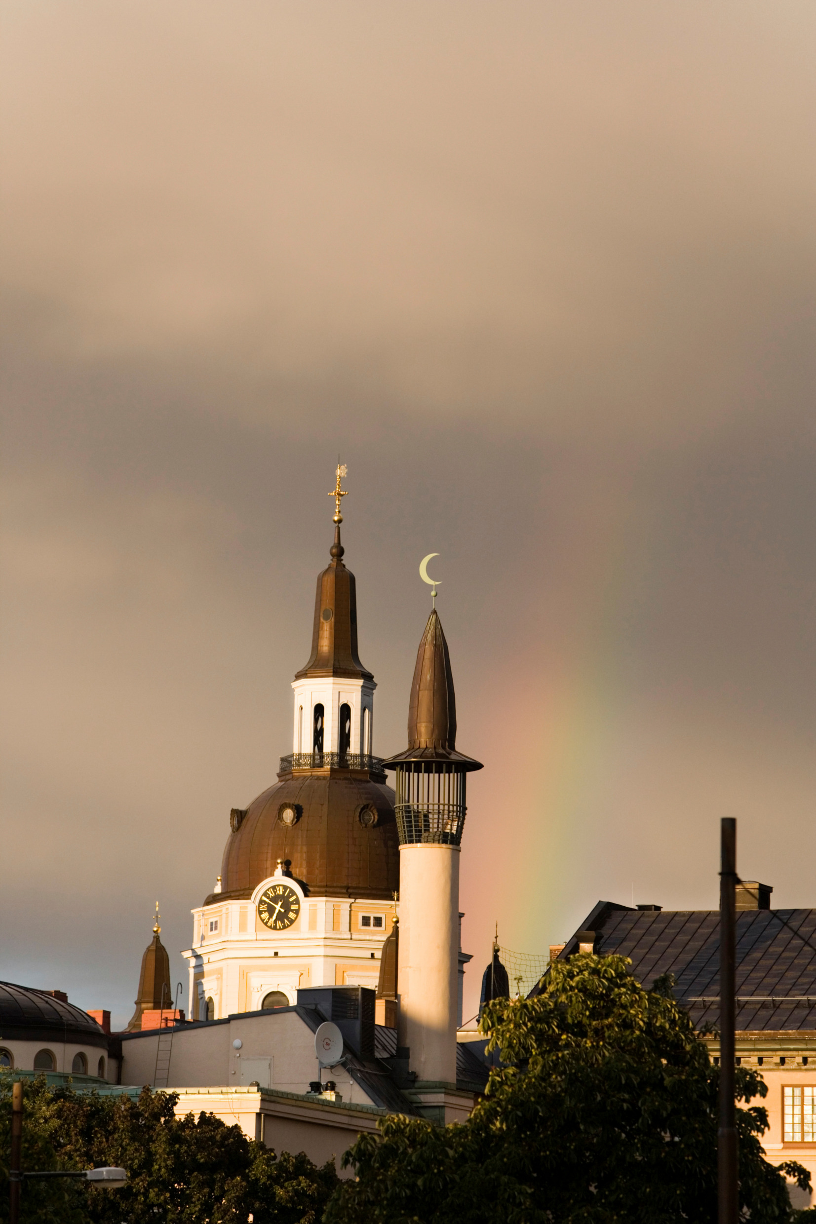 The tower of a church and a mosque against a dark sky with a rainbow.