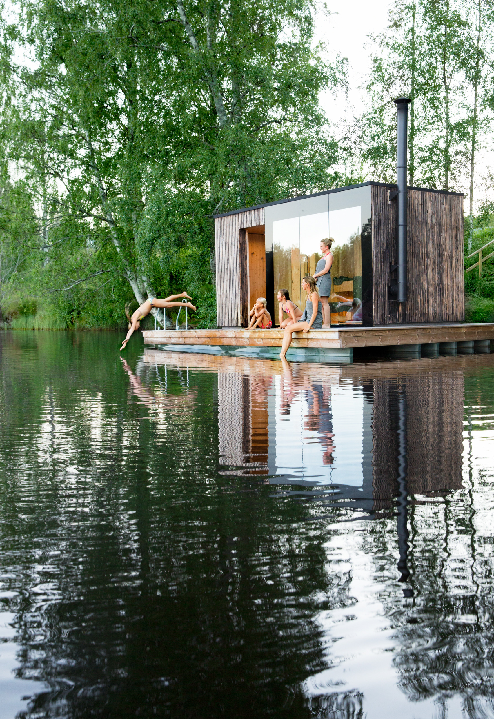 Agroup of women and girls in and around a sauna built on a small wooden pier by a lake.